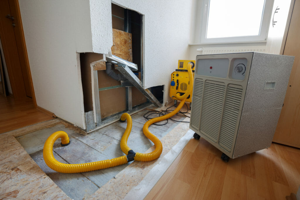 Gelber Bautrockner zum Wasserschaden beseitigen steht vor aufgestemmter Wand, gelbe Schläuche gehen in den Boden