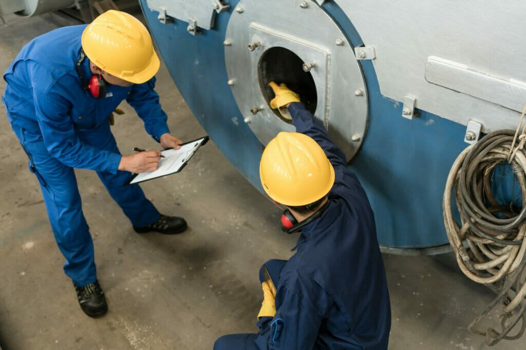 Zwei Arbeiter in blauer Arbeitskleidung und gelben Helmen ueberpruefen einen Thermaloelkessel auf seine Funktionstuechtigkeit