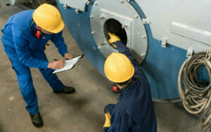 Zwei Arbeiter in blauer Arbeitskleidung und gelben Helmen ueberpruefen einen Thermaloelkessel auf seine Funktionstuechtigkeit