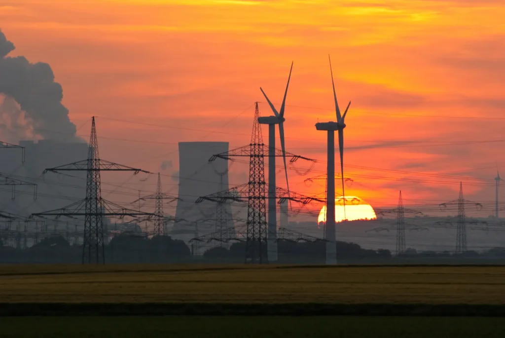 Windraeder stehen bei Sonnenuntergang vor Kohlekraftwerk und speisen Strom mit 50 Hz Netzfrequenz ins oeffentliche Stromnetz